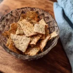 A glass bowl of crispy homemade seed crackers resting on a rustic wooden board beside a blue linen napkin.