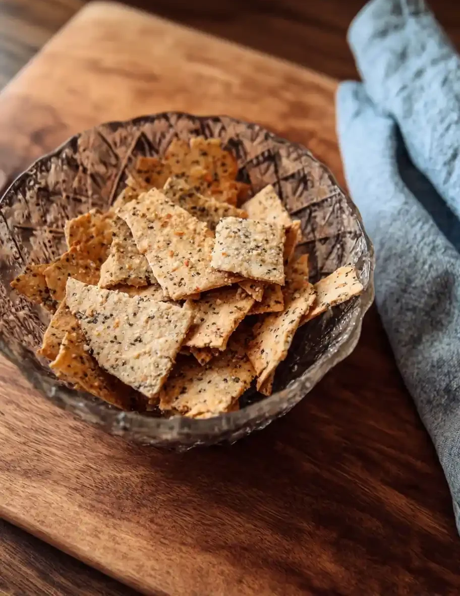 A glass bowl of crispy homemade seed crackers resting on a rustic wooden board beside a blue linen napkin.