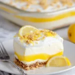 A creamy slice of layered lemon dessert on a white plate, garnished with fresh lemon zest and nuts.