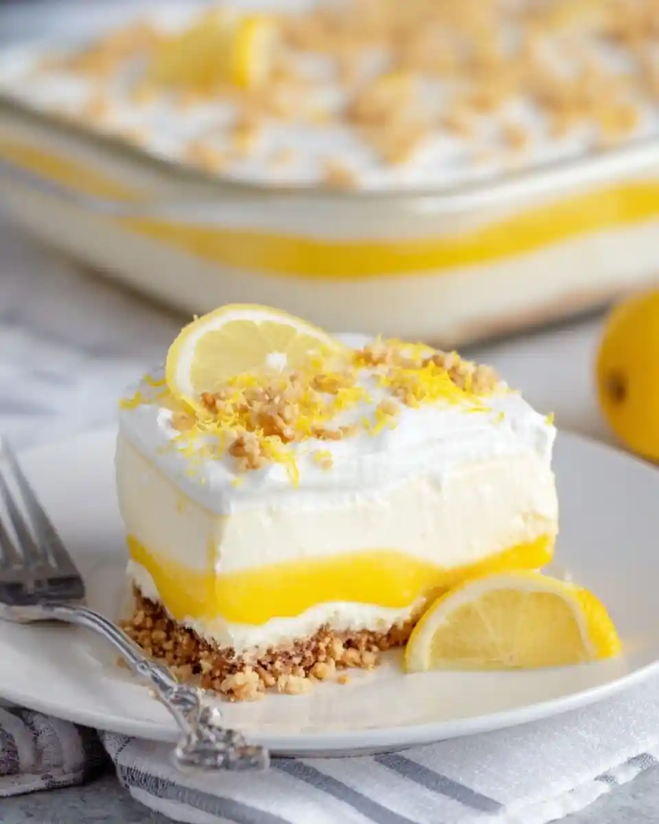 A creamy slice of layered lemon dessert on a white plate, garnished with fresh lemon zest and nuts.