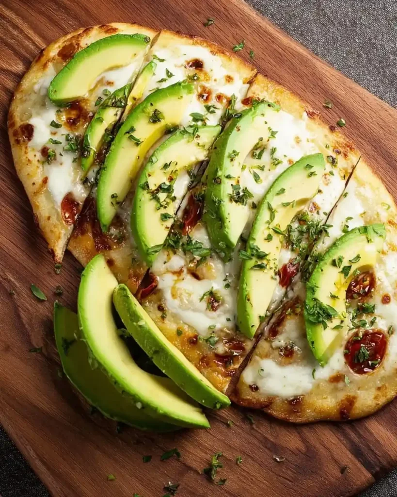 Freshly baked avocado flatbread with melted cheese, tomatoes, and herbs served on a rustic wooden board.