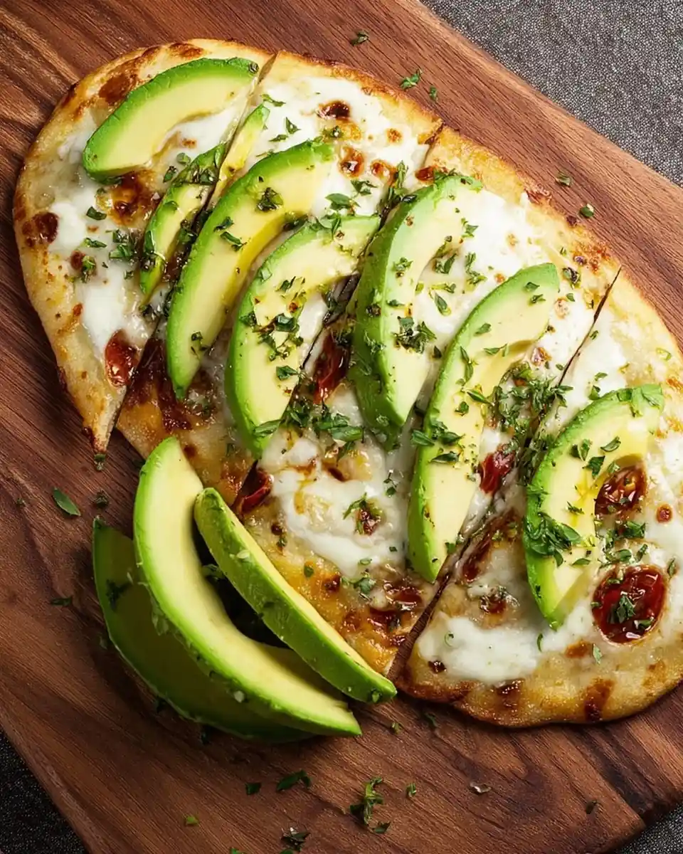 Freshly baked avocado flatbread with melted cheese, tomatoes, and herbs served on a rustic wooden board.