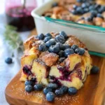 A slice of blueberry French toast casserole topped with fresh berries and powdered sugar on a wooden board.