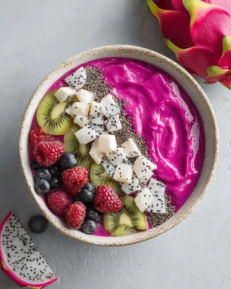 A bright pink dragon fruit smoothie bowl topped with kiwi, raspberries, blueberries, chia seeds, and diced dragon fruit.