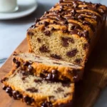 Sliced chocolate chip banana bread topped with a chocolate drizzle and chocolate chips on a wooden cutting board.