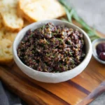 A white bowl of freshly made olive tapenade on a wooden board, accompanied by toasted baguette slices.