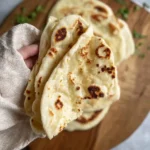 A hand holds a warm, buttery piece of naan bread with golden blistered spots over a wooden board.