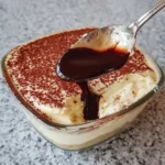 A spoon pouring chocolate syrup over a creamy {{ $('Fetch Recipe Data').first().json["Main Keyword"] }} dusted with cocoa.