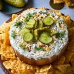 A wooden bowl filled with creamy dill pickle dip topped with pickles and dill, surrounded by crackers.
