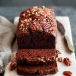 A freshly baked, sliced chocolate pecan loaf on a white cutting board, surrounded by scattered pecans.