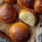 Fresh golden homemade hamburger buns with sesame seeds on parchment paper, with one cut open to show the soft interior.
