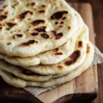 A stack of warm, fluffy {{ $('Fetch Recipe Data').first().json["Main Keyword"] }} with golden browned spots on a wooden board.