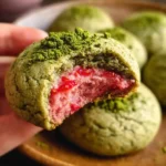 A hand holds a bitten treat, showcasing the gooey pink center of freshly baked matcha raspberry cookies.